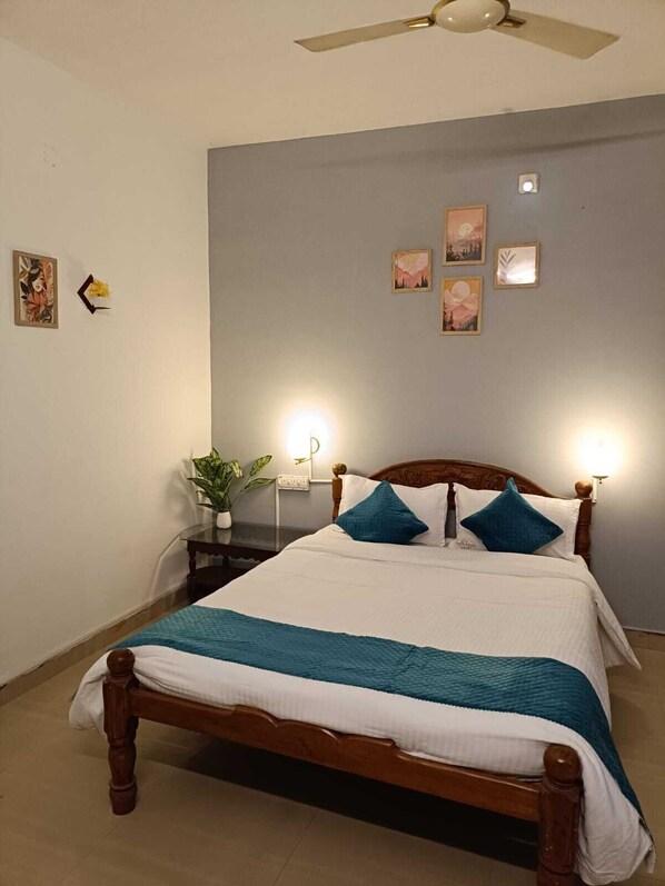 A neatly made bed with a wooden frame and white bedding, a small bedside table with a lamp, and a ceiling fan. - Villa De Grand (Kuilapalayam)