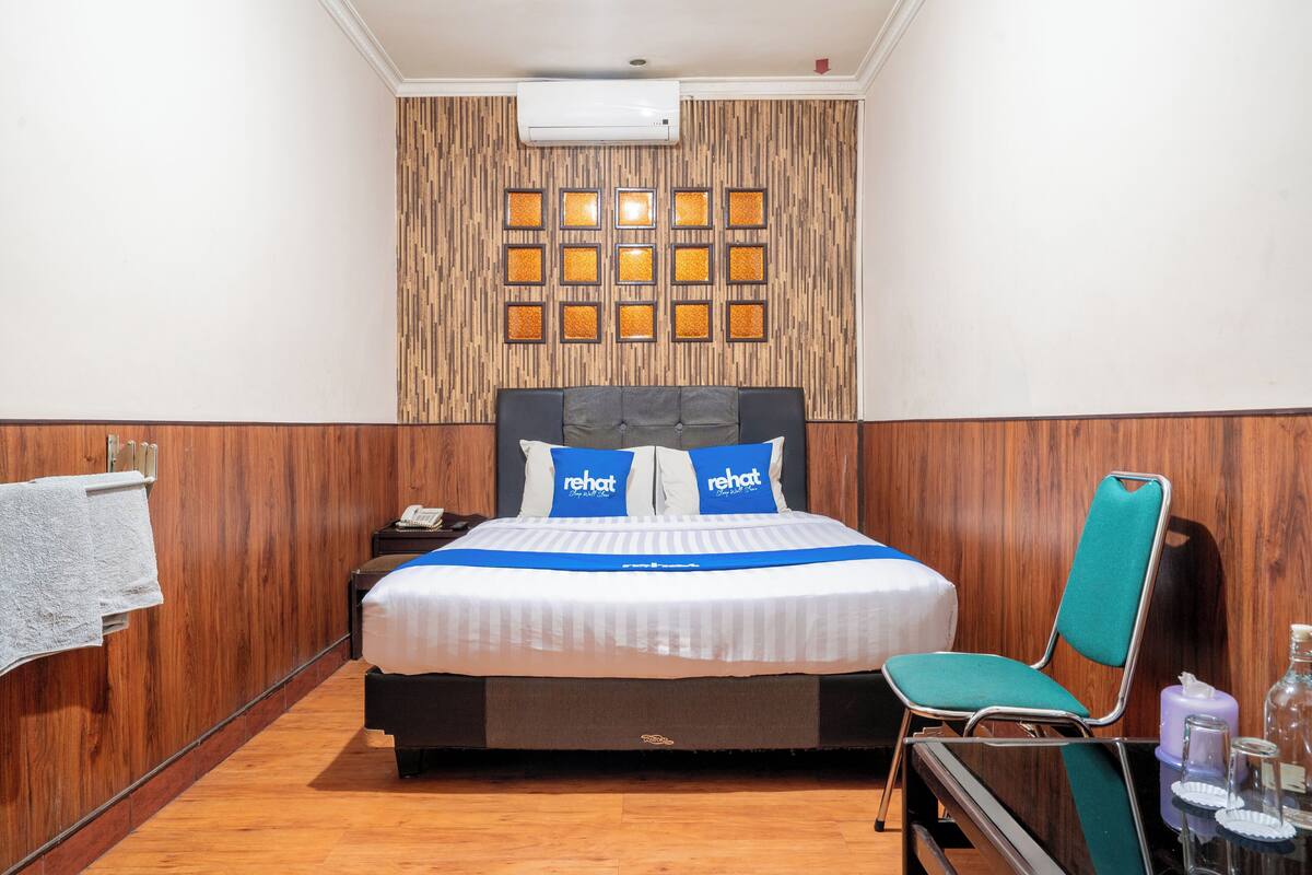 A hotel room with a bed, a chair, a nightstand, a wall-mounted air conditioner, and a wooden panel wall with framed pictures.