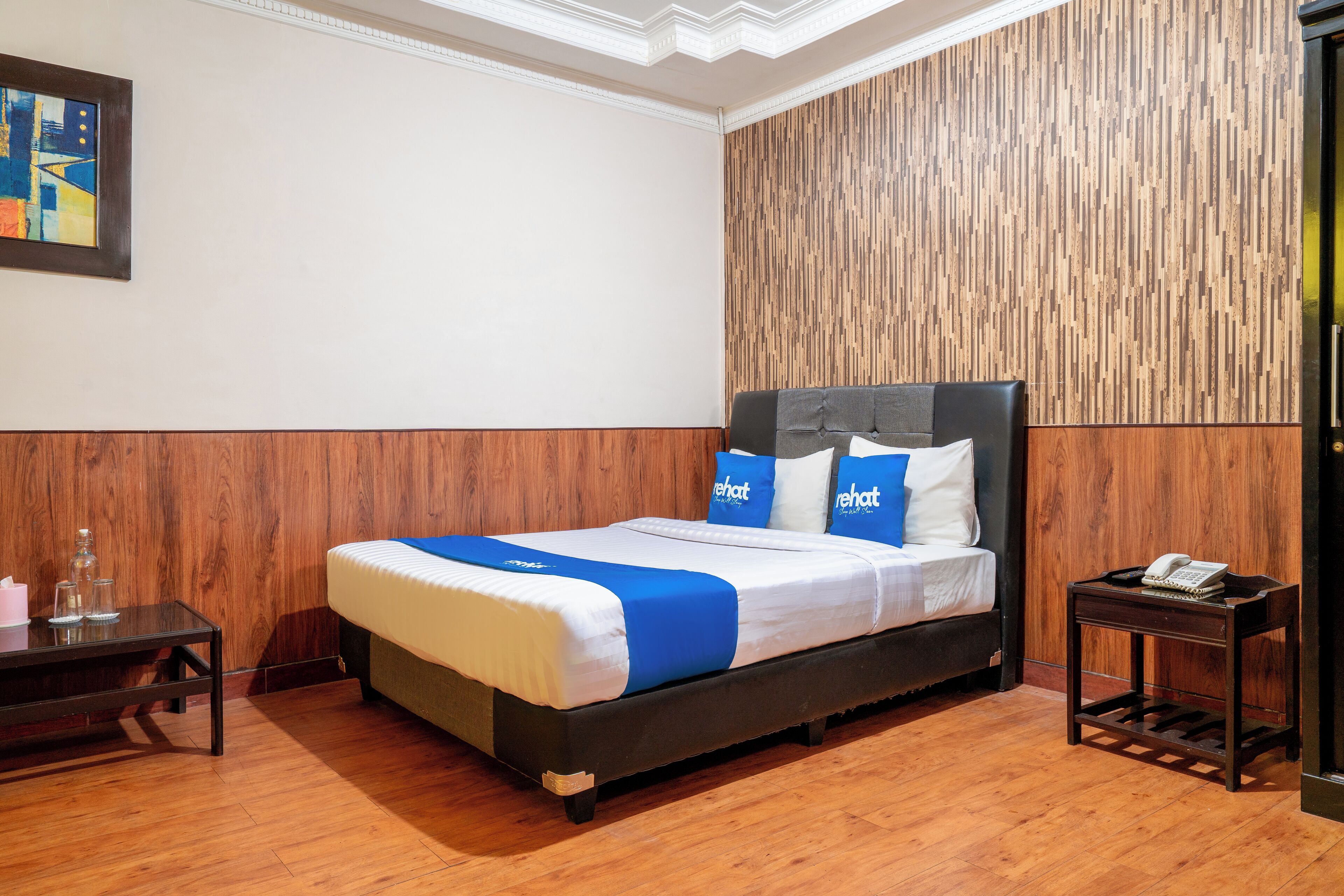 A hotel room with a wooden floor, a bed with blue and white bedding, a small wooden nightstand, and a wooden accent wall.