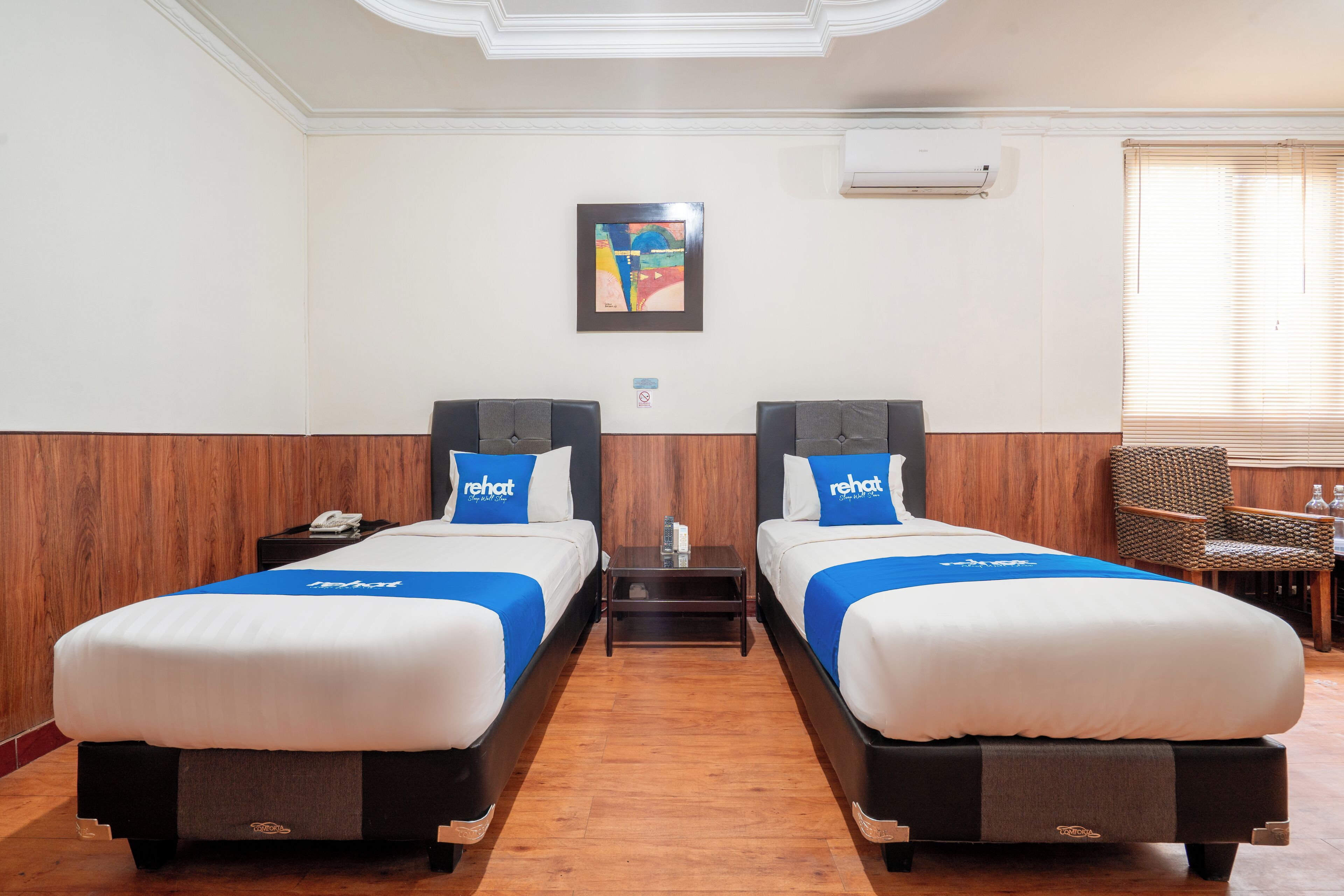 A hotel room with two beds, each with a blue headboard and matching pillow, a small table with a phone and a bottle, and a framed abstract painting on the wall.