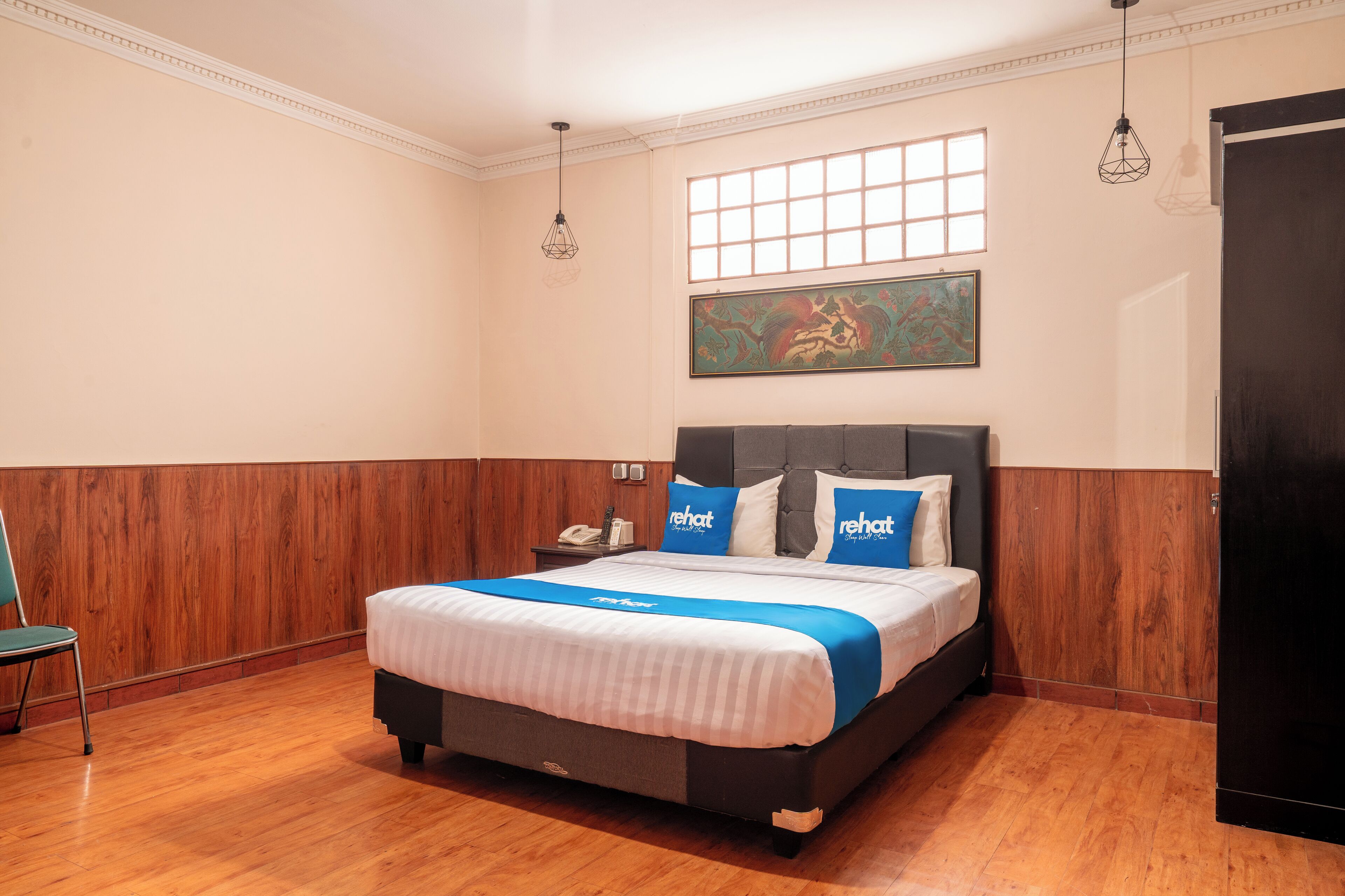A hotel room with a wooden floor, a bed with a blue and white bedspread, a gray headboard, and a painting on the wall.