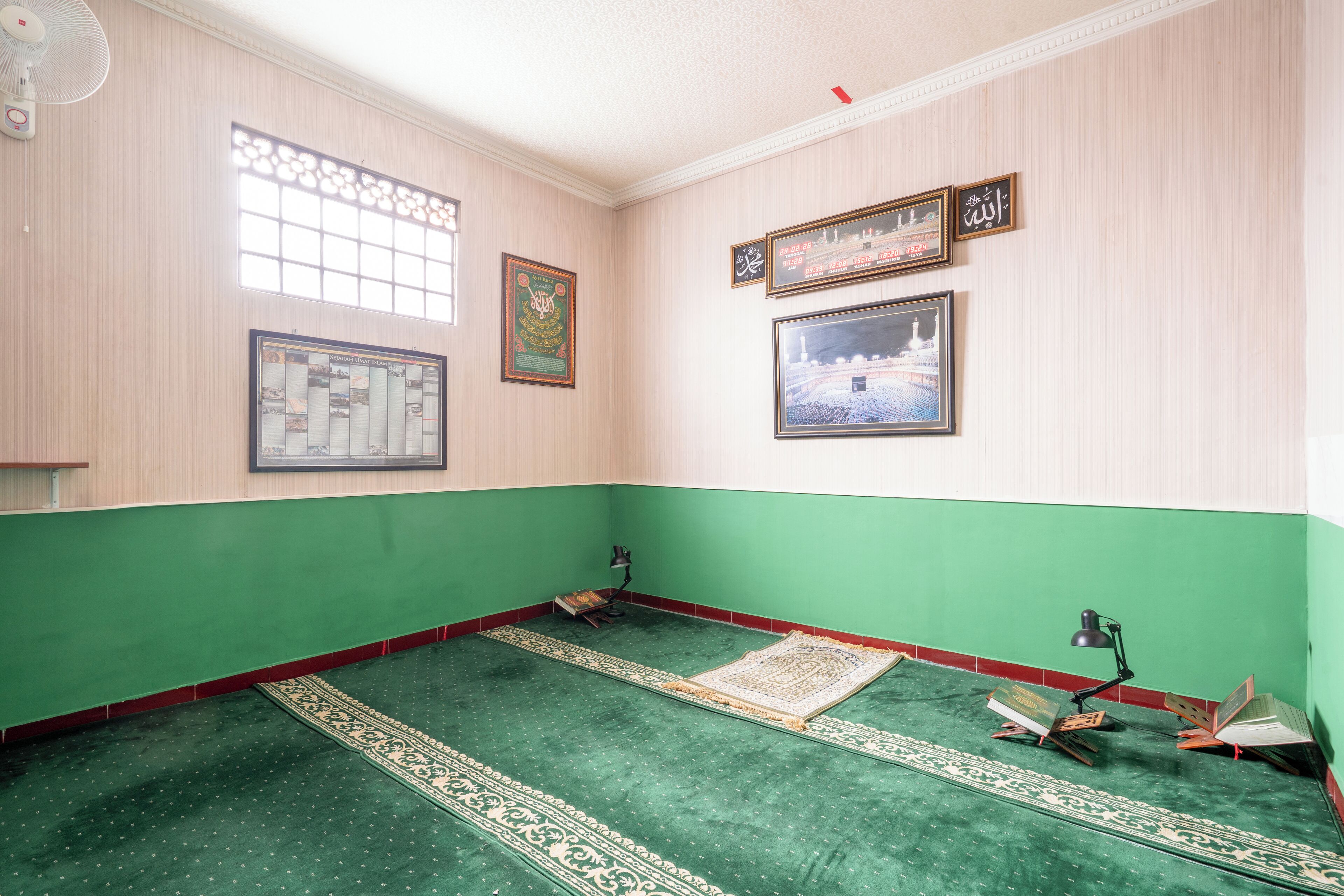 A prayer room with green carpet, prayer mats, a prayer rug, a bookshelf, and framed religious texts on the wall.