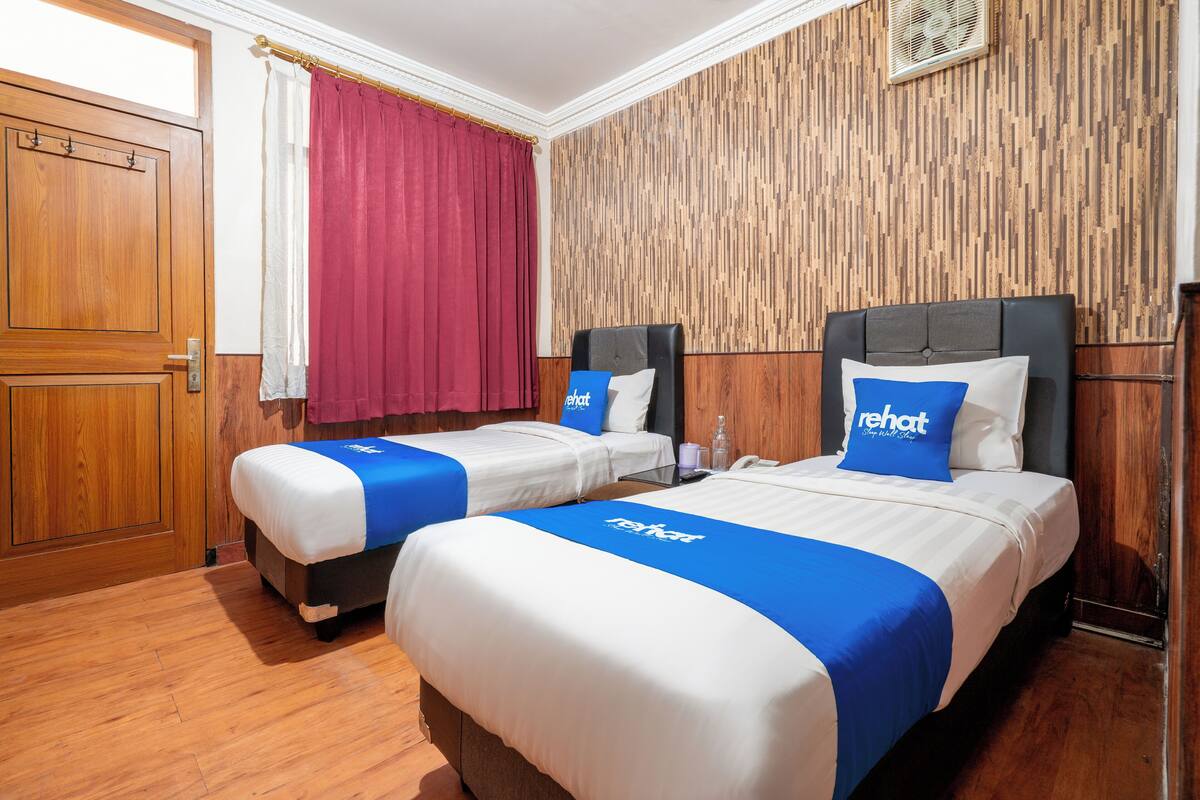 A hotel room with two beds, wooden flooring, a wooden wardrobe, a red curtain, and a wall with a vertical wood panel design.