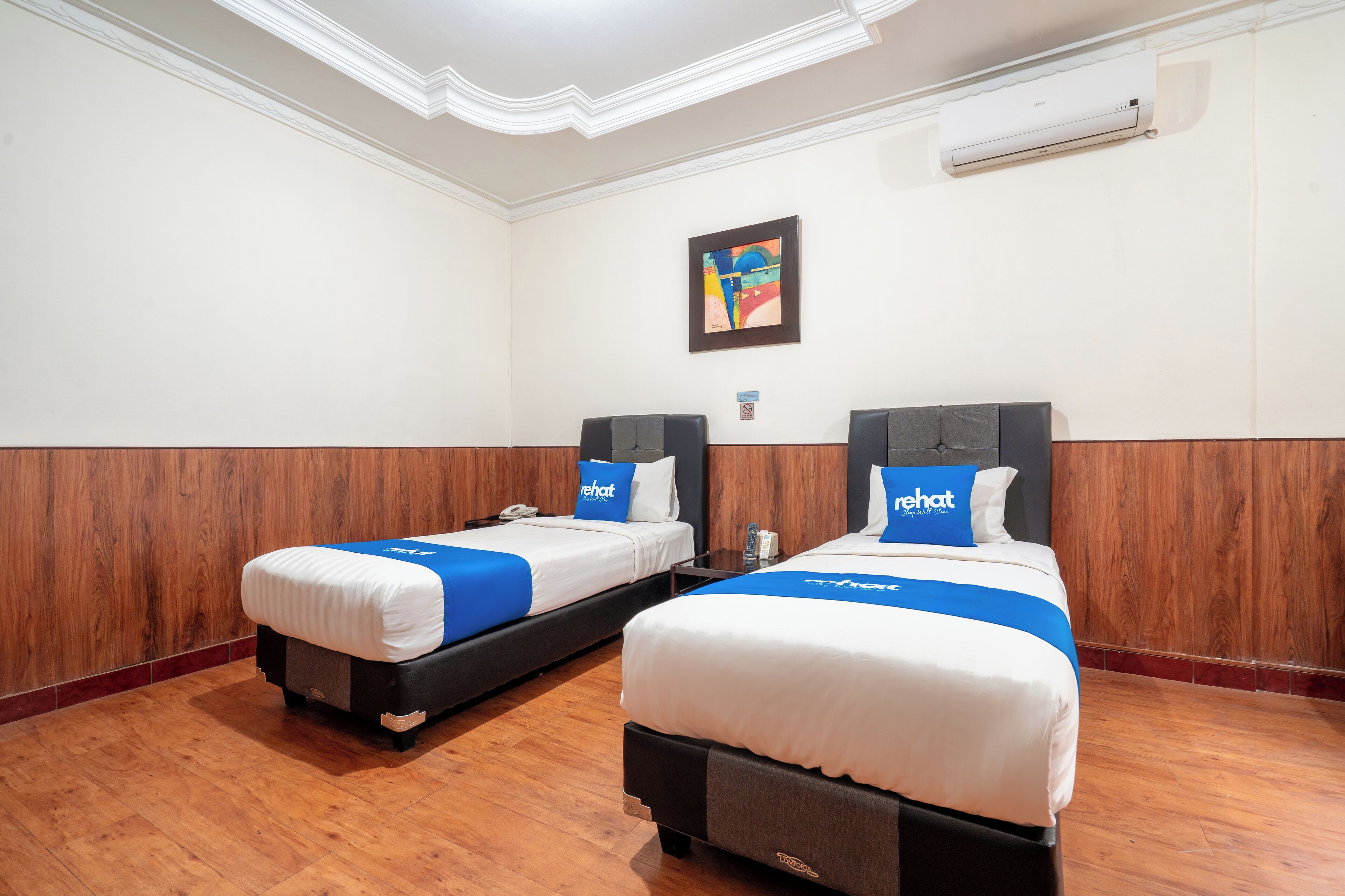 Twee eenpersoonsbedden met hoofdborden, een houten vloer, een witte muur met een airconditioning en een lijst met een afbeelding boven de bedden.