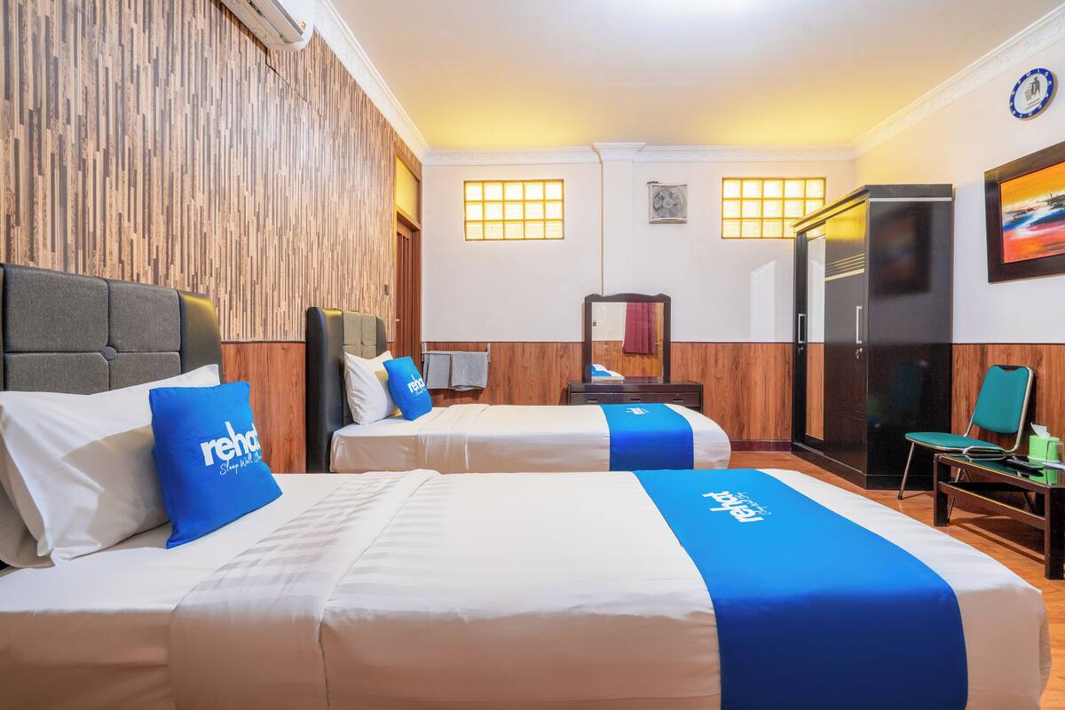 A hotel room with two beds, wooden paneling on the walls, a refrigerator, a chair, and a clock on the wall.