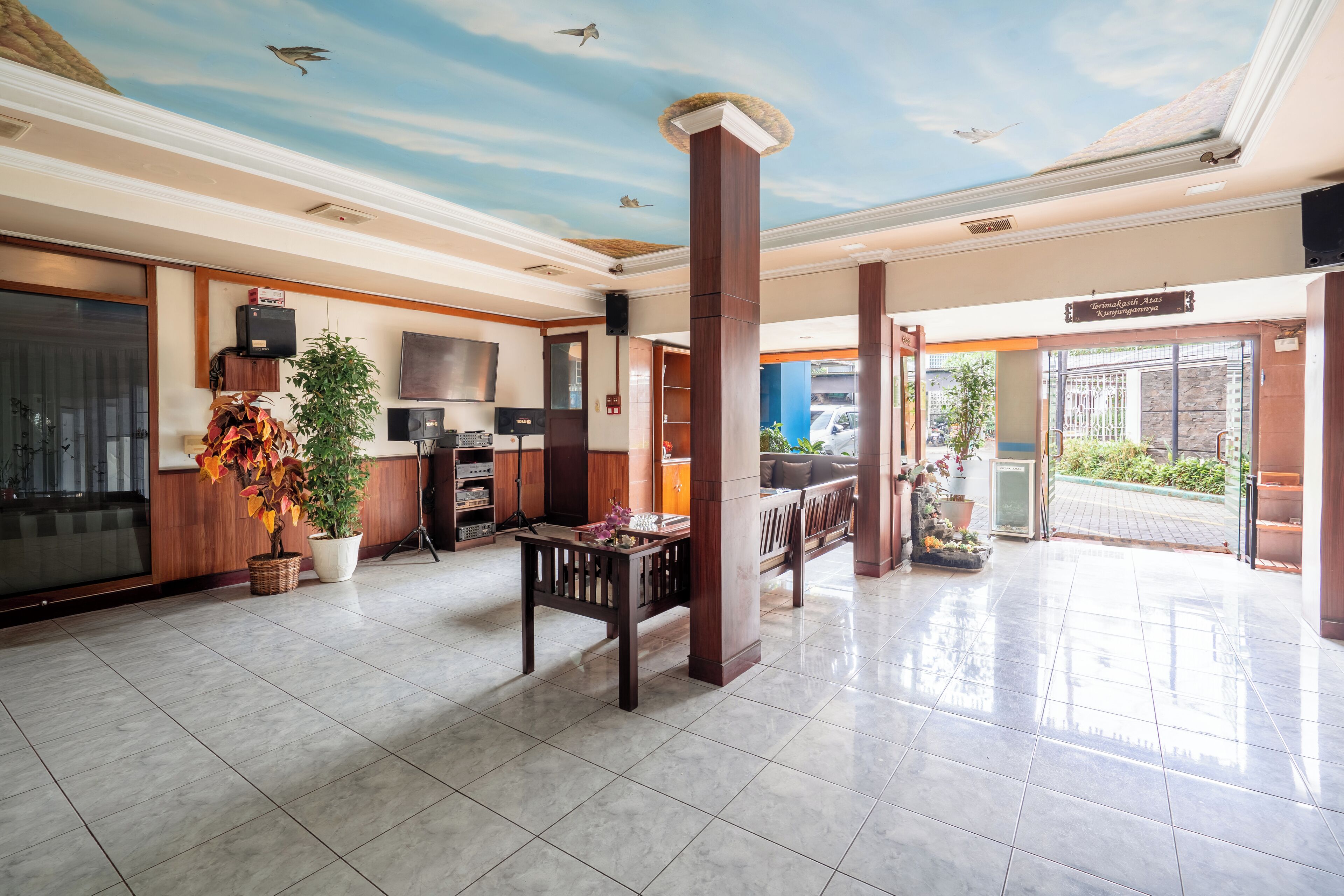 A spacious lobby with a tiled floor, wooden furniture, a large potted plant, and a ceiling mural.