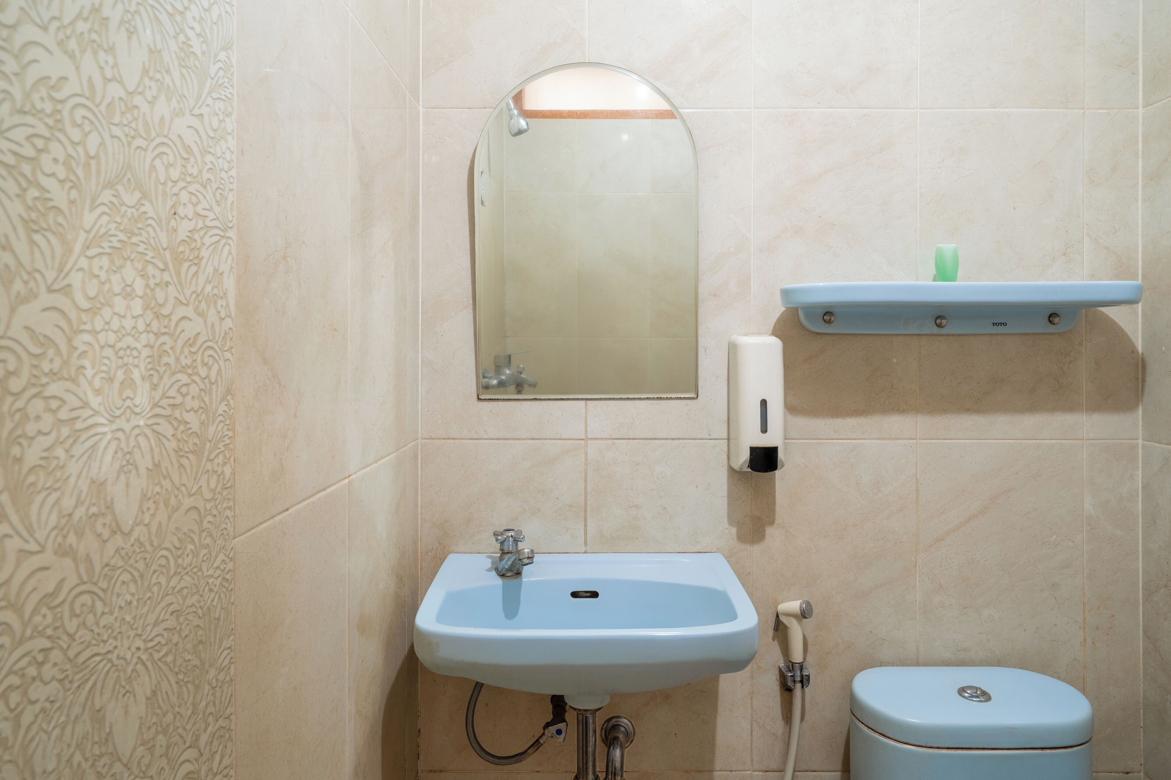 A bathroom with a sink, mirror, toilet, and a soap dispenser on a shelf.