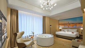 Een hotelkamer met een groot bed, een rond ligbad, een zithoek met een stoel en tafel, en uitzicht op de stad.