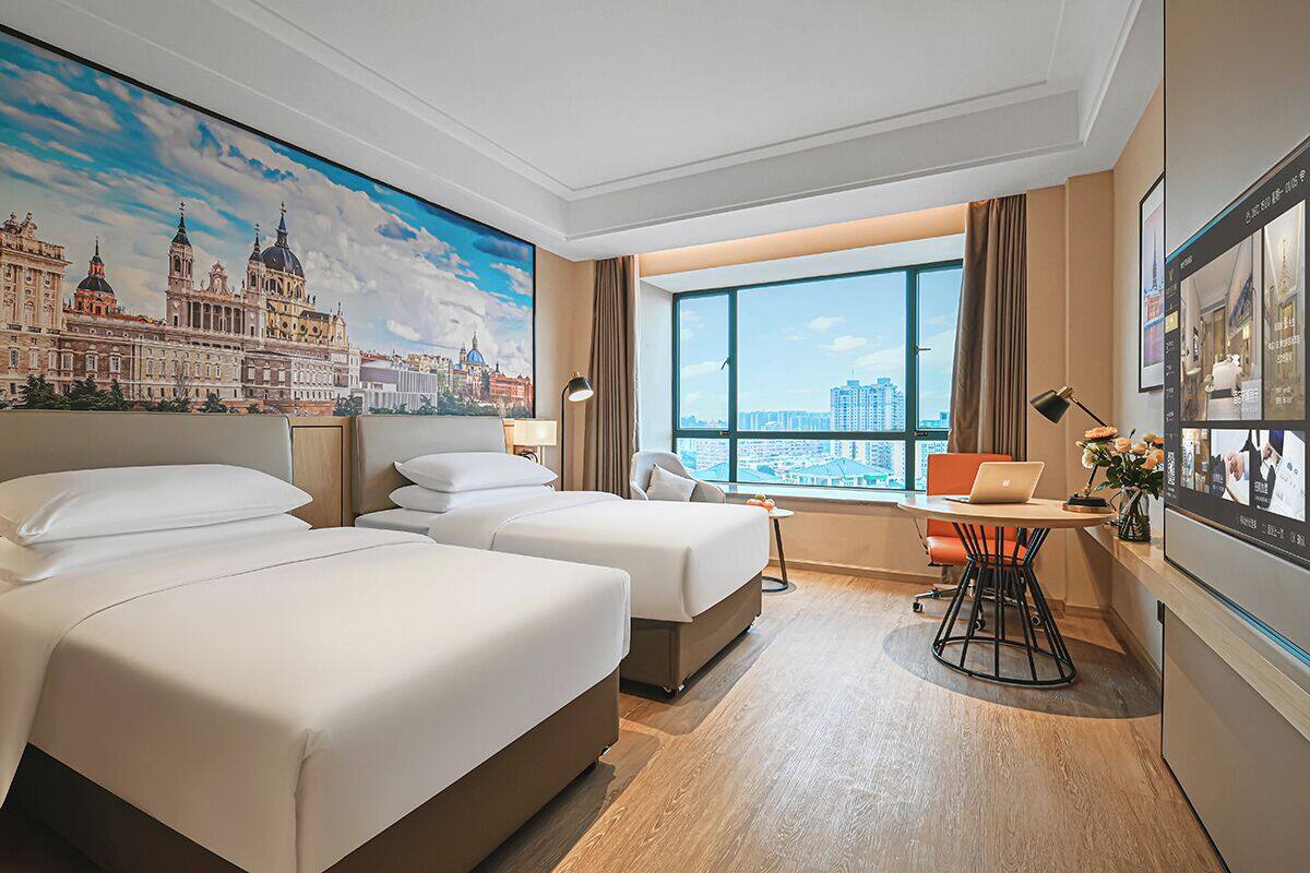 Ein Hotelzimmer mit zwei Betten, einem Schreibtisch mit einem Laptop, einem großen Fenster mit Blick auf die Stadt und einem Gemälde eines historischen Gebäudes.