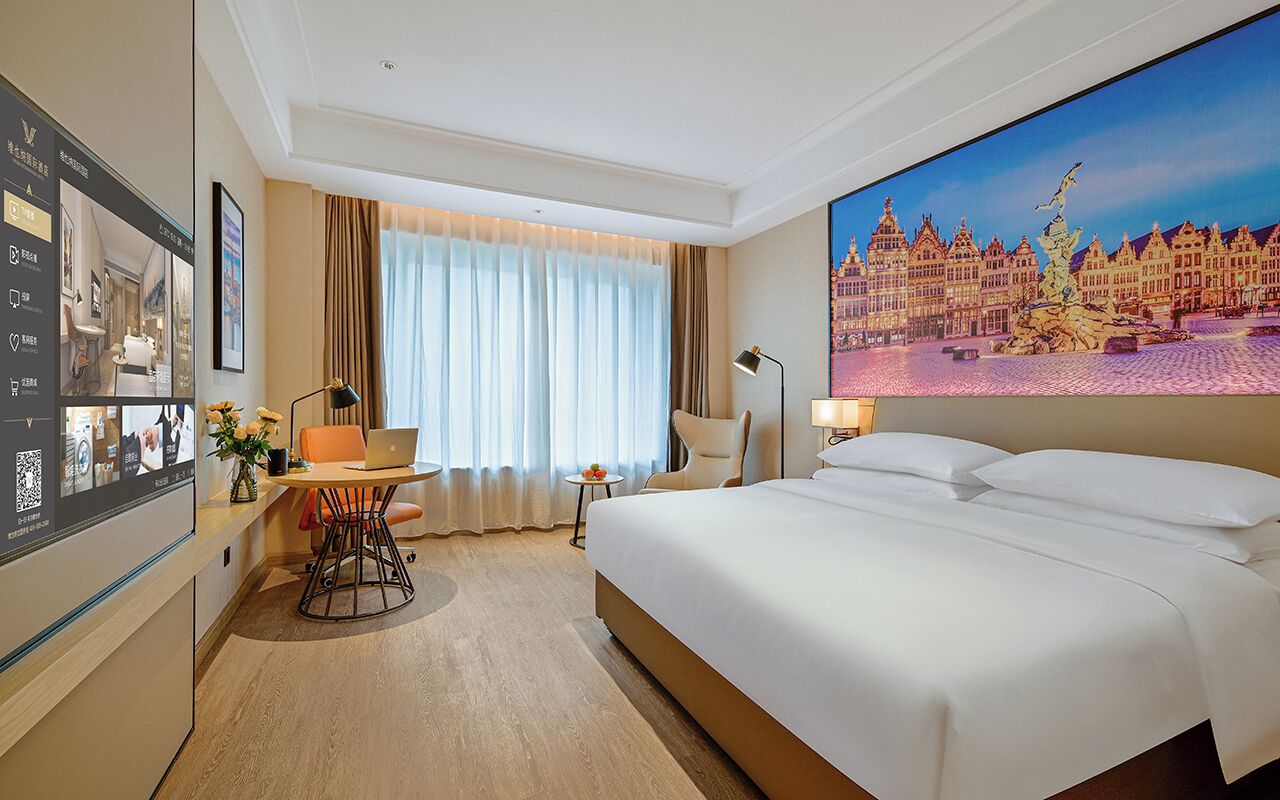 A hotel room with a bed, a desk with a laptop, a chair, a small table with fruit, a lamp, and a large window with curtains.