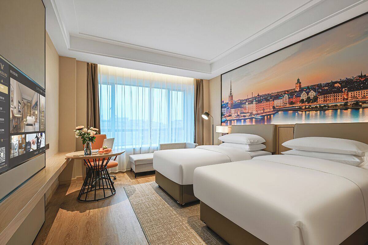 Ein Hotelzimmer mit zwei Betten, einem Schreibtisch mit einem Laptop, einem Fenster mit Vorhängen und einem Stadtbild an der Wand.