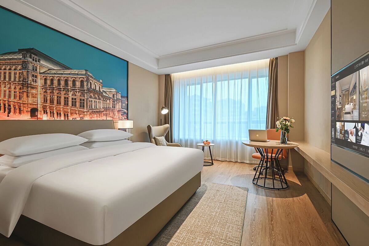 Ein Hotelzimmer mit einem großen Bett, einem Schreibtisch mit einem Laptop, einem Sessel, einem kleinen Tisch mit Blumen und einem Bild eines Gebäudes an der Wand.