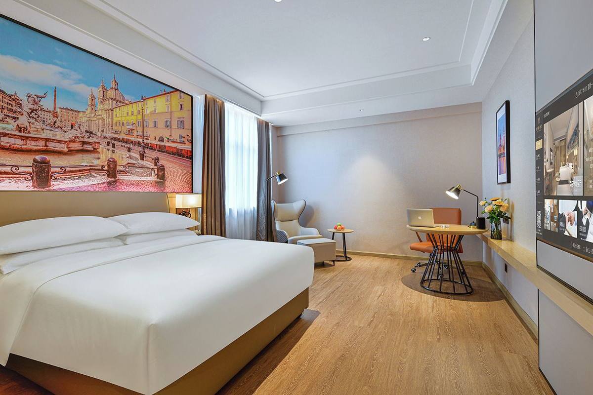 Ein Hotelzimmer mit einem großen Bett, einem Schreibtisch mit Stuhl, einem kleinen Tisch mit Lampe und einem Stadtbild an der Wand.