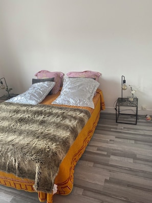 A bed with a patterned blanket and pillows, a small bedside table with a lamp, and a wall socket. - Large 4-bedroom house with garden - near D-Day beaches and train station (Sainte-Marguerite-d'Elle)