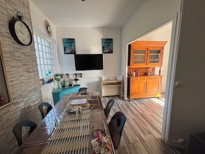 A dining area with a long table, chairs, a clock on the wall, and a kitchen in the background. - Large 4-bedroom house with garden - near D-Day beaches and train station (Sainte-Marguerite-d'Elle)