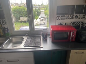 A kitchen with a sink, a red microwave, and a window with a view of greenery outside. - Large 4-bedroom house with garden - near D-Day beaches and train station (Sainte-Marguerite-d'Elle)