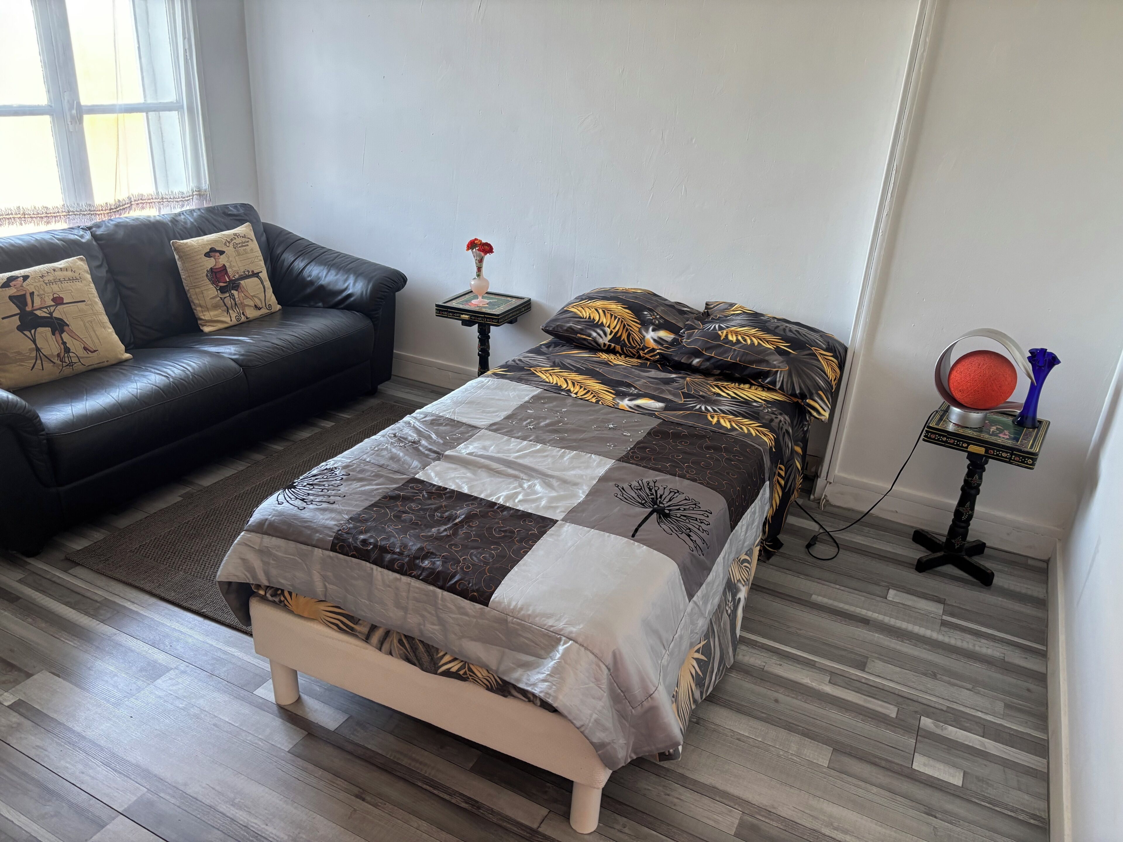 A bedroom with a bed, a sofa, a small table with a vase, and a decorative item on a stand.