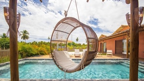 A hanging chair over a swimming pool with a view of a house and palm trees. - Casa com Piscina e Quadra no Condom nio By Toque (São Miguel dos Milagres)