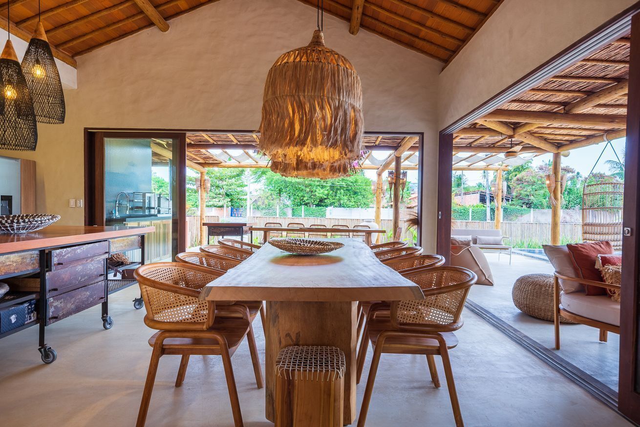 A spacious dining area with a large wooden table and wicker chairs, a hanging light fixture, and a view of the outdoors.