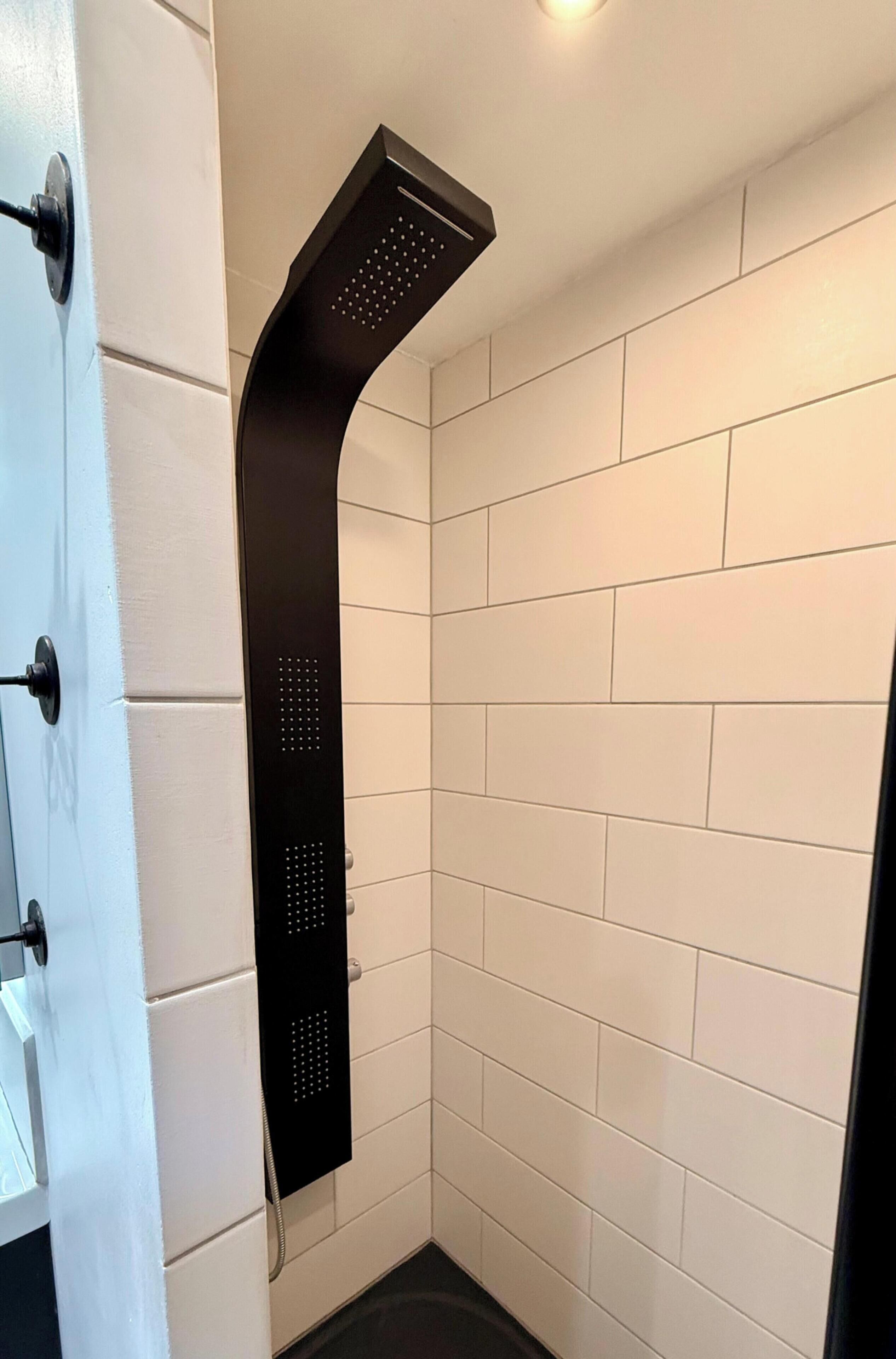 A modern bathroom with white tiled walls, a black showerhead, and a visible shower control panel.