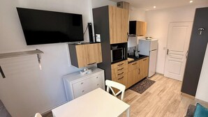 A compact kitchen with a flat-screen TV, wooden cabinets, a white countertop, and a small dining area. - Alpine Sanctuary by the Market Lane (Mont-Dore)