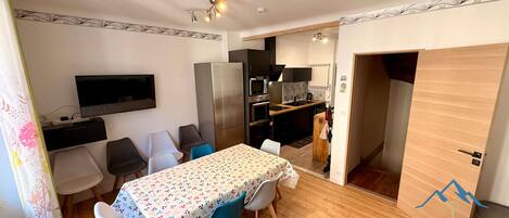 Espaço compacto com área de refeições, cozinha compacta e uma pequena TV fixada na parede.