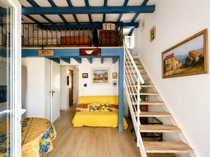 A room with a wooden staircase, a yellow sofa, a bed with floral bedding, and a painting on the wall. - Charming and Quiet Cottage – Sainte-Maxime (STE MAXIME)