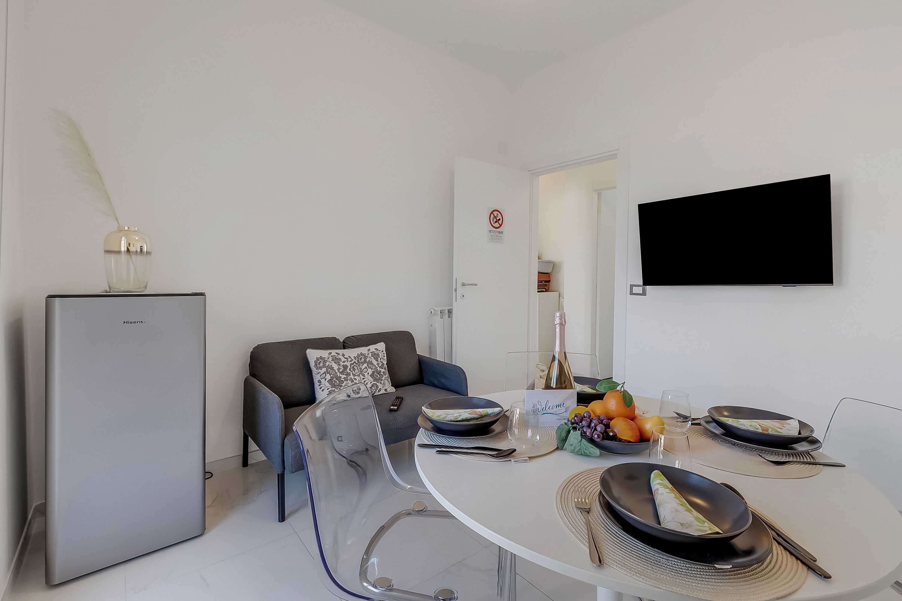 A modern dining area with a round table, clear chairs, a flat-screen TV, a refrigerator, and a sofa with a decorative pillow.