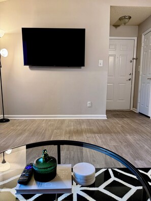 A living room with a flat-screen TV mounted on the wall, a floor lamp, a coffee table with a decorative item, a remote control, and a stack of coasters. - Quiet & Cozy Nook, Prime Location Near Everything! (Atlanta)