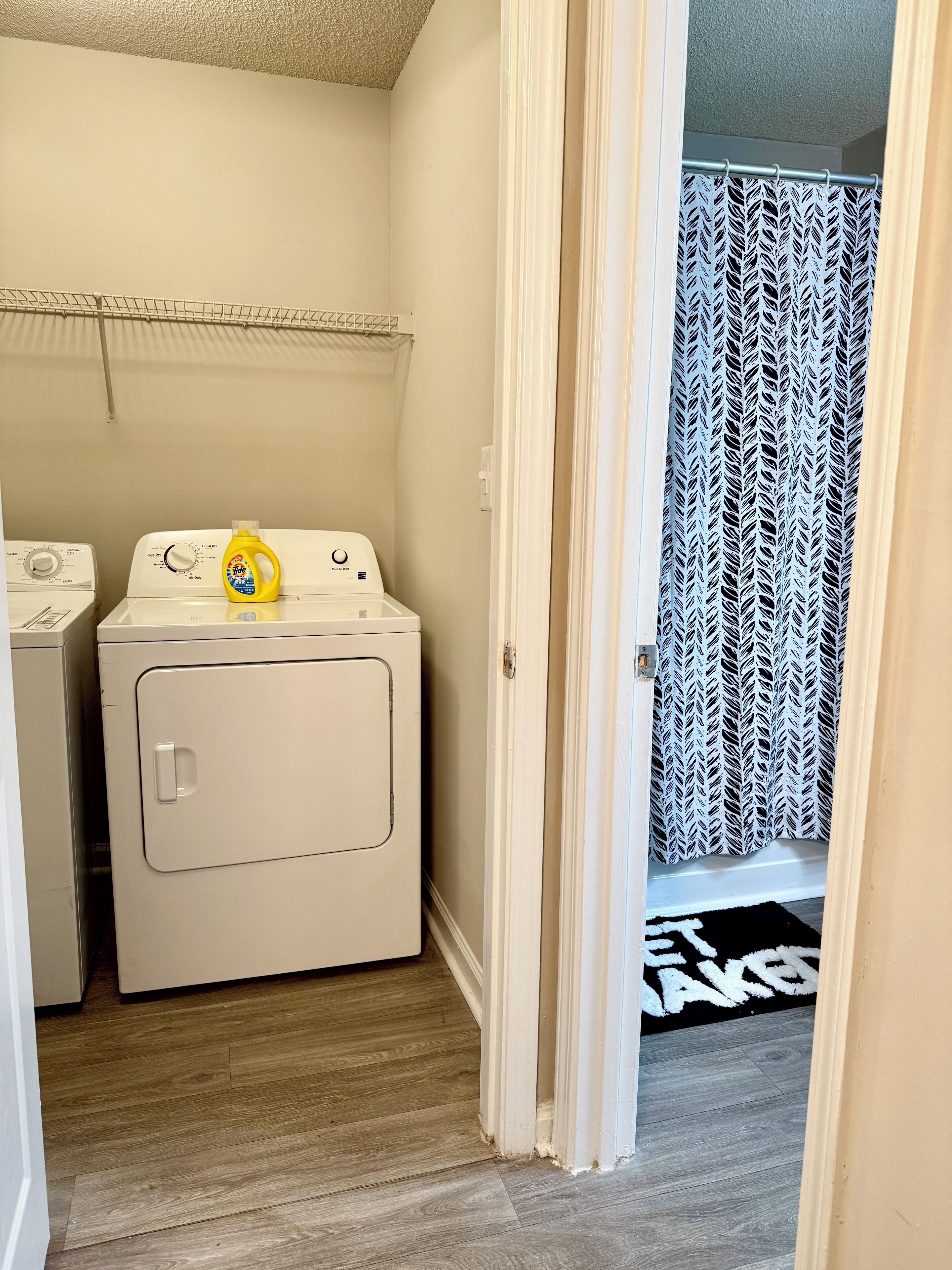 A laundry room with two washing machines, a yellow detergent bottle on the dryer, and a patterned shower curtain in the adjacent bathroom.