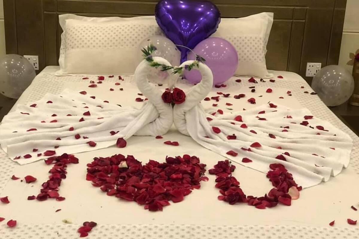 A bed with rose petals arranged in a heart shape, a bouquet of roses, and heart-shaped balloons.