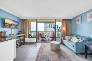 A modern living room with a blue sofa, a coffee table, and a TV on the wall. There is a balcony with red chairs and a view of buildings. - Cozy oceanfront 1-bedroom condo in beautiful Myrtle Beach (Myrtle Beach)