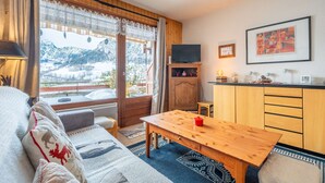A cozy living room with a wooden table, a sofa with patterned cushions, a TV, and a kitchen area with wooden cabinets. - Loges A : Grande terrasse à 50m des pistes. (LE GRAND BORNAND)