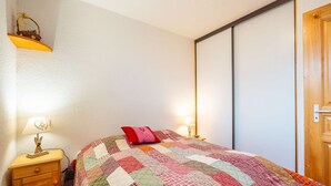 A bedroom with a quilted bedspread, a bedside lamp, a wooden nightstand, and a wooden door. - Loges A : Grande terrasse à 50m des pistes. (LE GRAND BORNAND)
