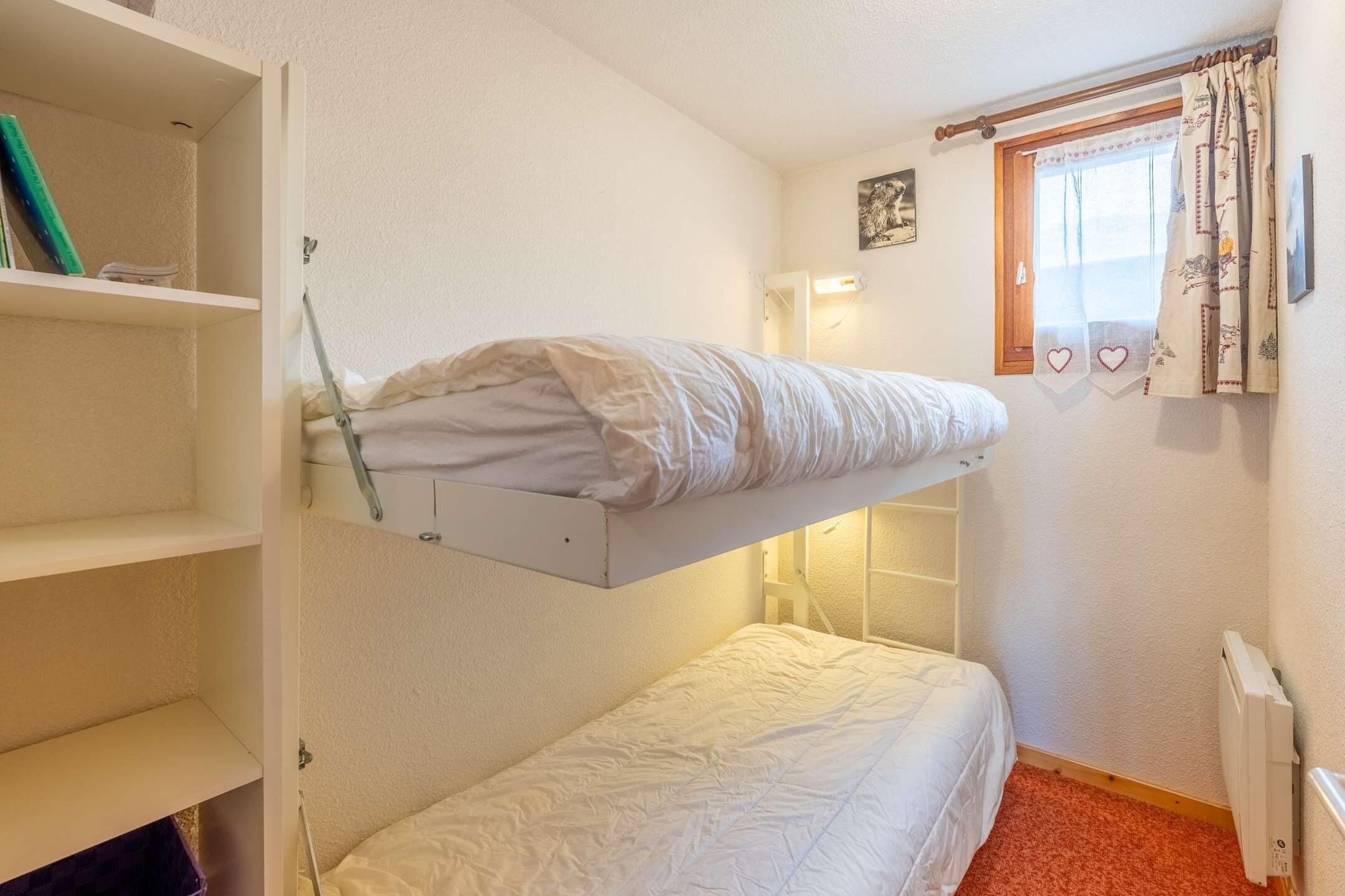 Una cama litera con colchón blanco, estante con libros, radiador y ventana con cortinas.