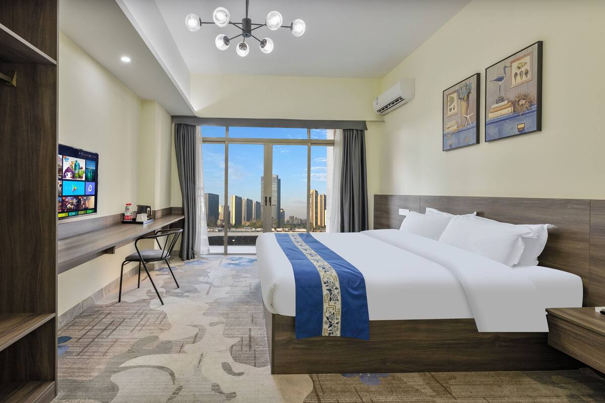 A hotel room with a large bed, a desk with a chair, a TV, and a city view.