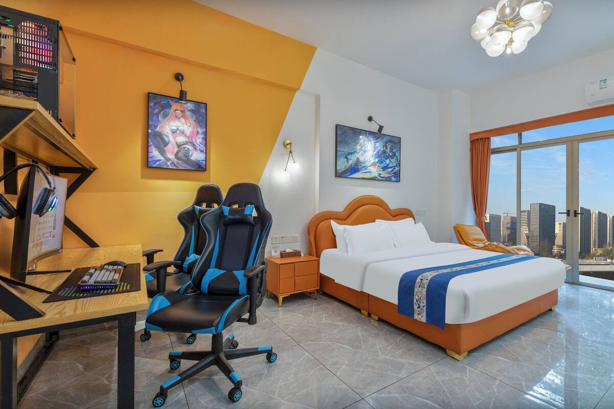 A hotel room with a bed, desk, gaming chair, and a large window with a city view.