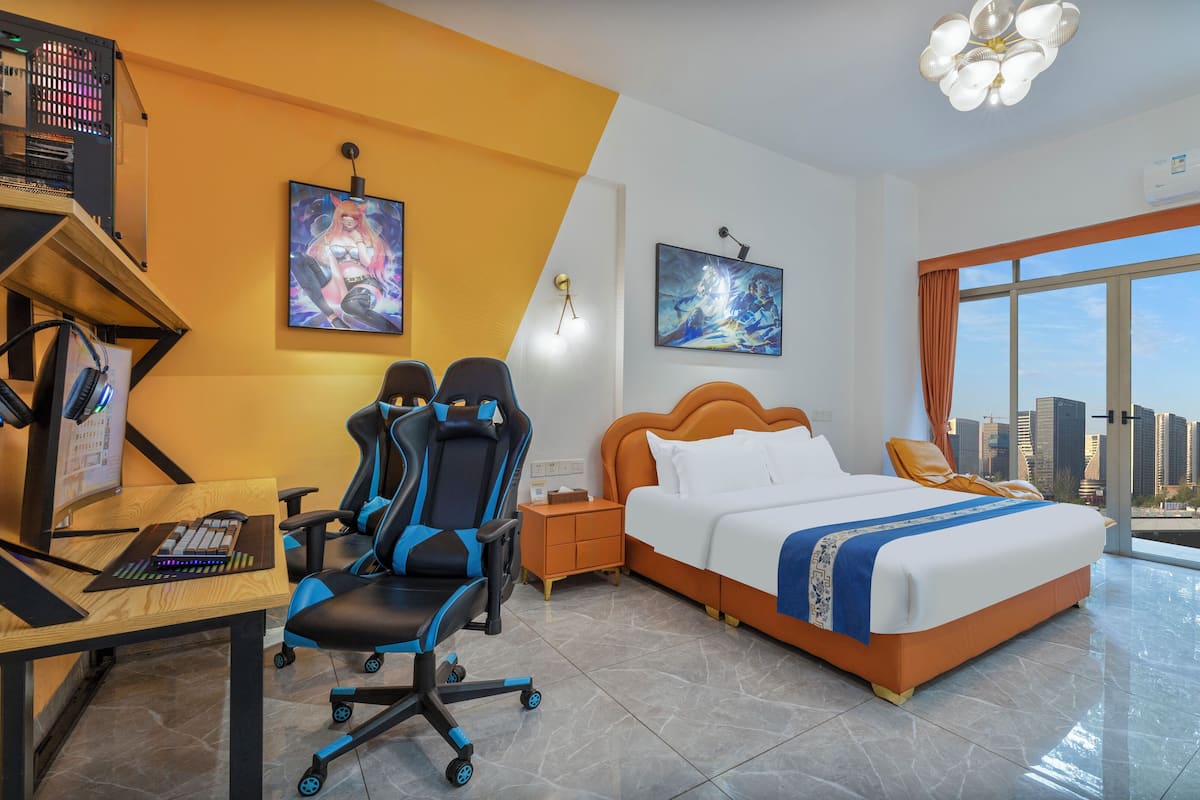 A hotel room with a bed, desk, gaming chair, and a large window with a city view.