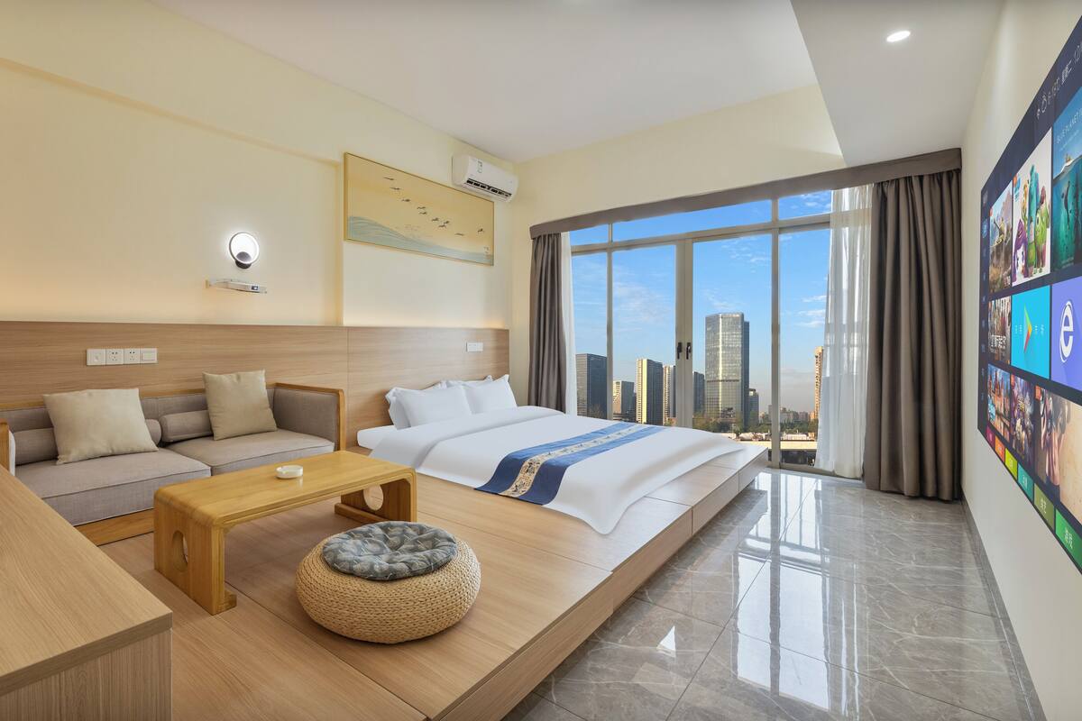 A modern hotel room with a bed, sofa, coffee table, and a large window with a city view.