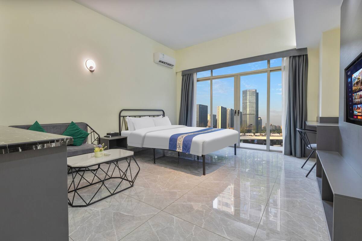 A modern hotel room with a bed, a desk, a chair, a small table, and a city view.
