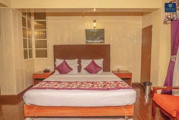 Quarto de hotel com uma cama grande, cabeceira de madeira, duas mesas de cabeceira, um criado-mudo, um cesto de lixo e uma cadeira.