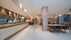 Un hall d’hôtel moderne comprenant un comptoir d’accueil, une horloge numérique, un téléviseur et un espace repas avec des tables et des chaises.