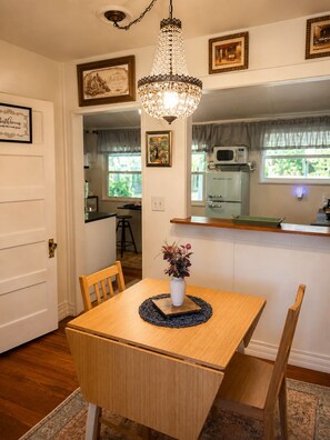 A dining area with a wooden table, chairs, a vase with flowers, a chandelier, and a kitchen in the background.