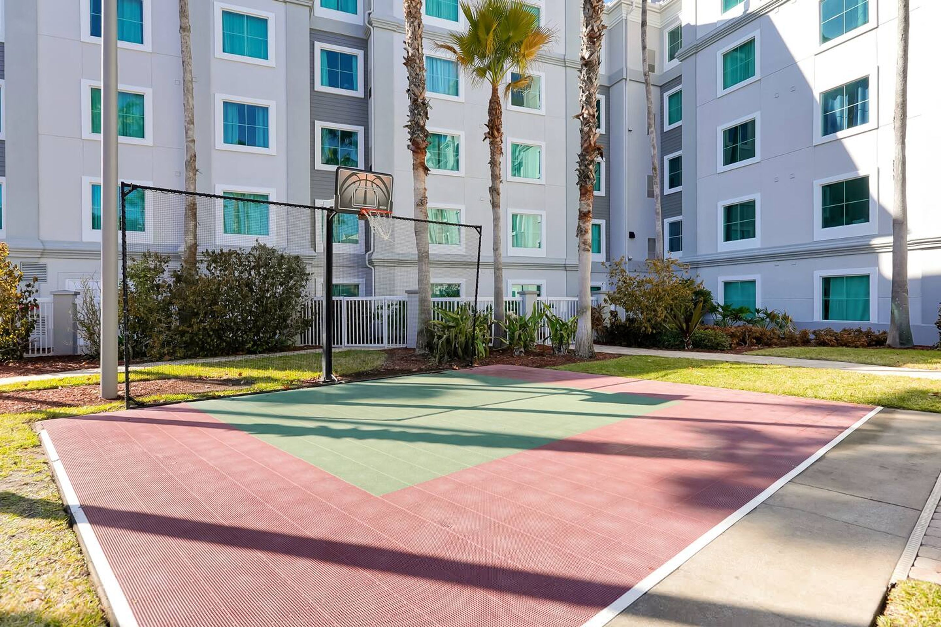 Ein Basketballplatz im Freien neben einem mehrstöckigen Gebäude mit weißer Fassade und grünen Fenstern.