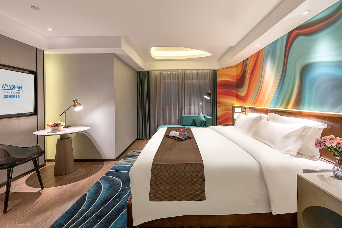 Ein Hotelzimmer mit Bett, Schreibtisch, Stuhl, Fernsehbildschirm und einem farbenfrohen Wandgemälde.
