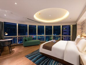 Una habitación de hotel moderna con una cama grande, un sofá verde, un escritorio con una silla y vistas a la ciudad de noche.