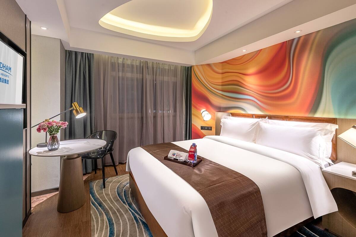Ein Hotelzimmer mit Bett, Nachttisch, Lampe und einem farbenfrohen, abstrakten Wandbild.
