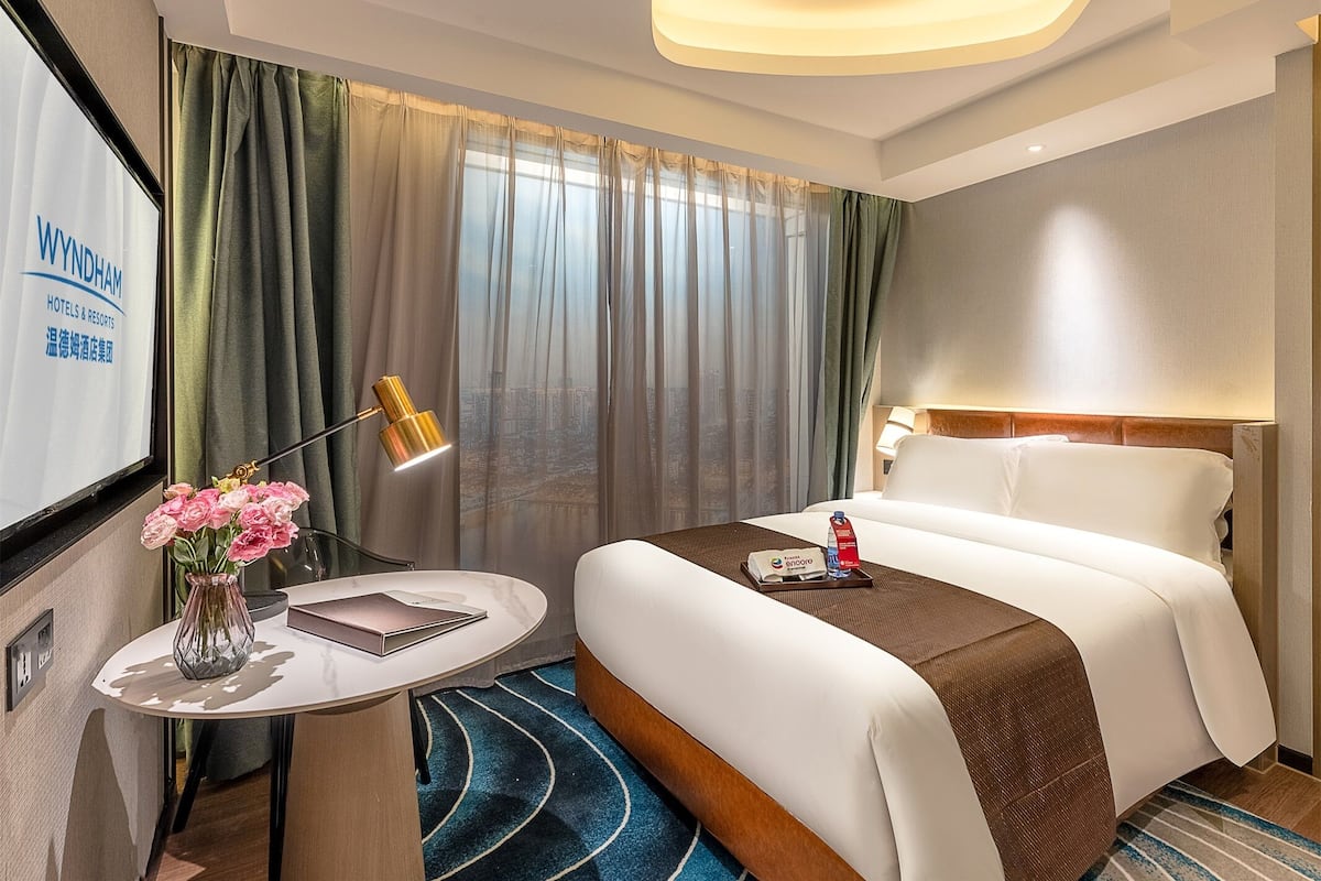 Ein Hotelzimmer mit Bett, Nachttisch mit Lampe, Blumenvase und Blick auf die Stadt durch das Fenster.