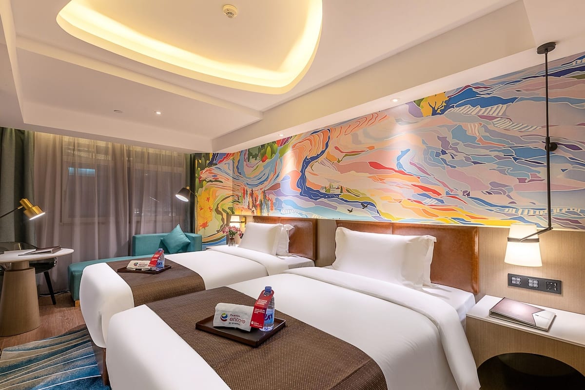 Ein Hotelzimmer mit zwei Betten, einem farbenfrohen Wandgemälde, einem Schreibtisch mit Lampe und Blick auf die Stadtlandschaft.