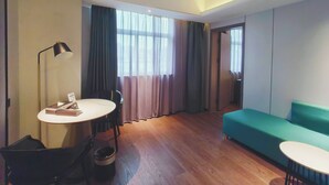 Moderni hotellihuone, jossa on puulattia, pyöreä ruokapöytä tuoleineen, vihreä sohva ja työpöytä lampin kanssa.